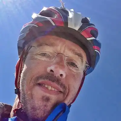 Fröhliche Portraitaufnahme vor blauem Himmel, leicht schräg von unten aufgenommen. Auf dem Kopf ein roter Radhelm mit weißen Streifen, im Gesicht etwas unrasiert und sonnenverbrannt.
